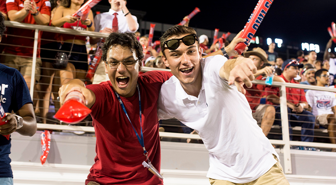 Two male students during an MŮ football game smiling and pointing at the camera