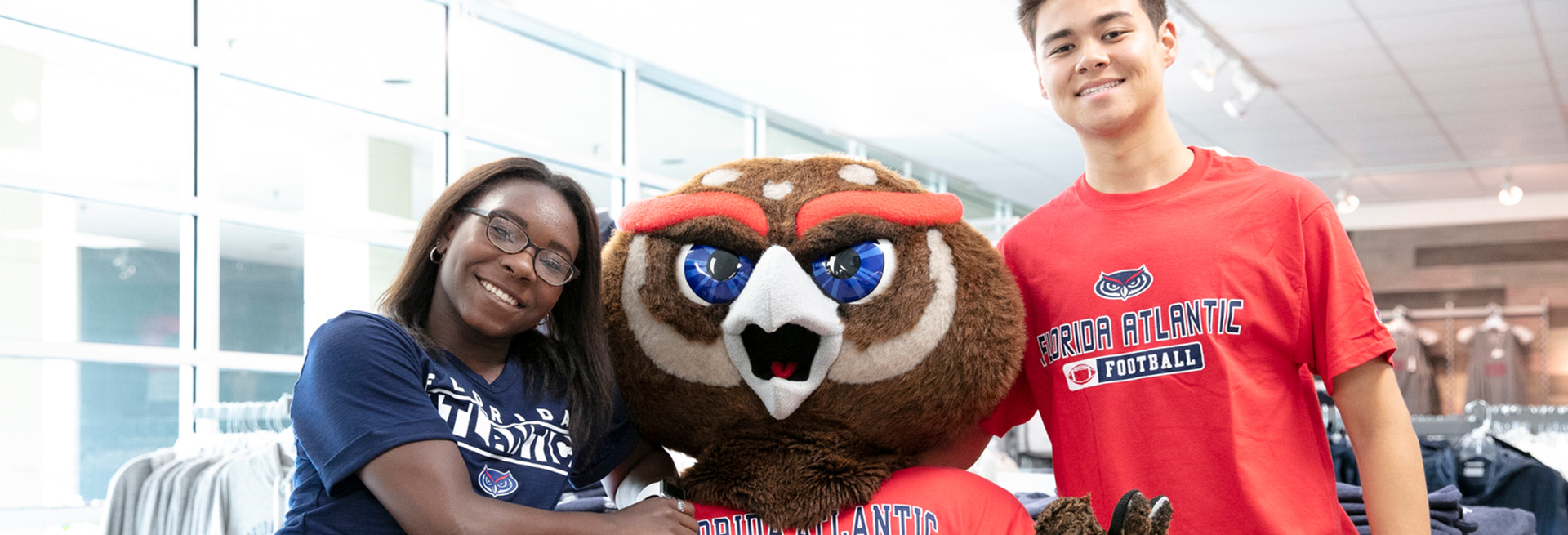 Two students next to MŮ mascot Owlsley wearing MŮ merchandise